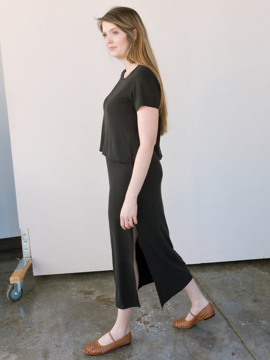 Black ribbed nursing dress with built-in bralette for breastfeeding. Modeled by a woman against a white background standing in a casual pose.