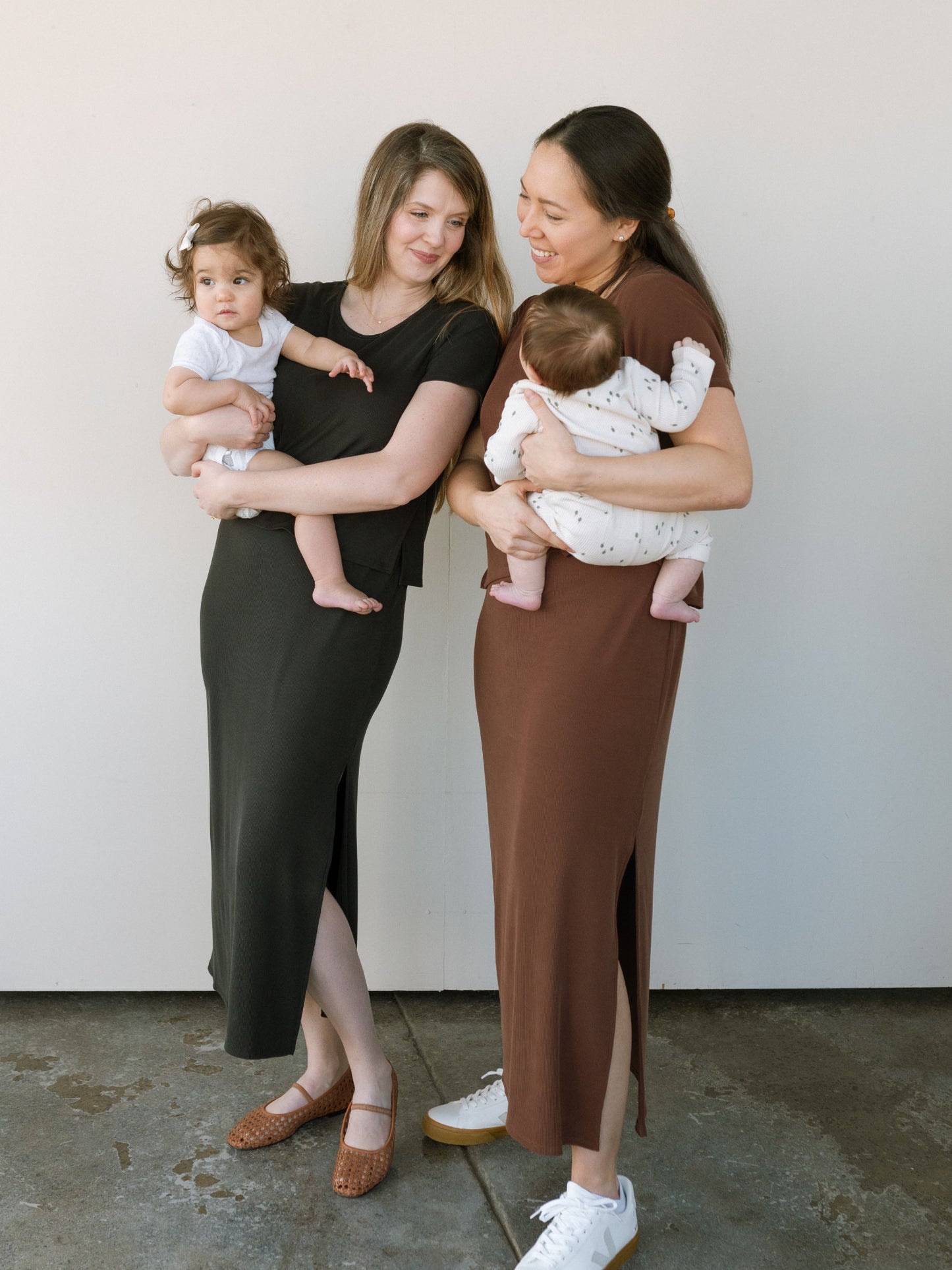 Two woman standing together, holding their babies and smiling. They wear their cute and comfortable nursing-friendly dresses, one in black and one in brown.