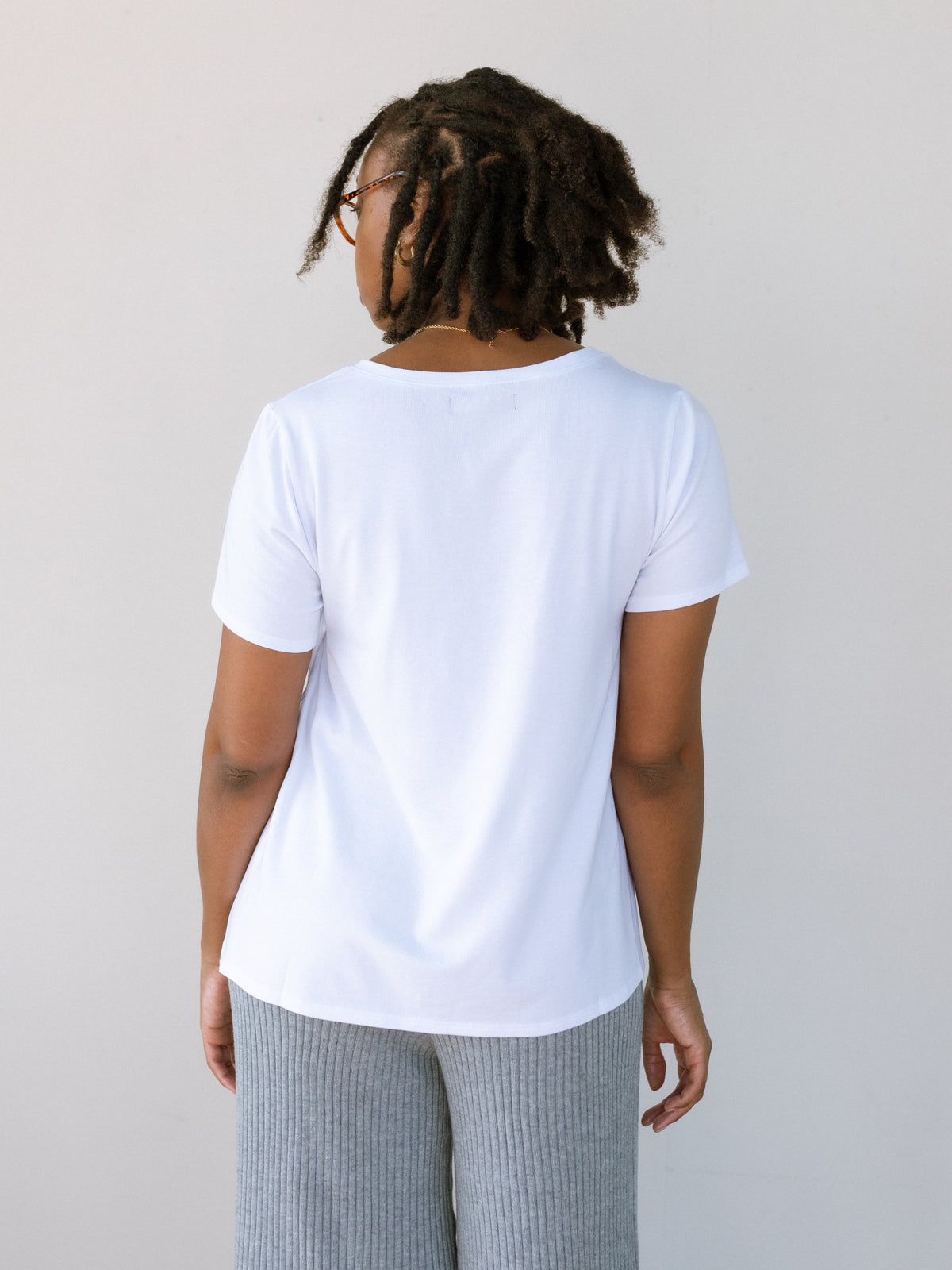 A woman standing against a white backdrop, showing the back of her white nursing tshirt.
